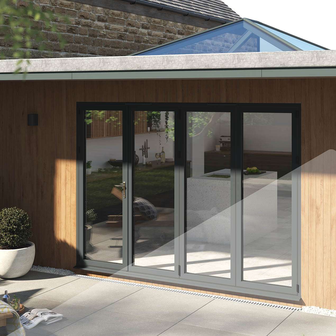 Exterior view of a wooden panelled flat roof extension with a set of grey Korniche bi-folding doors and a Korniche rood lantern installed.The patio outside has a plant pot situated to the left of the doors and in the bottom left corner a cocktail and an open book are visible on the ground.