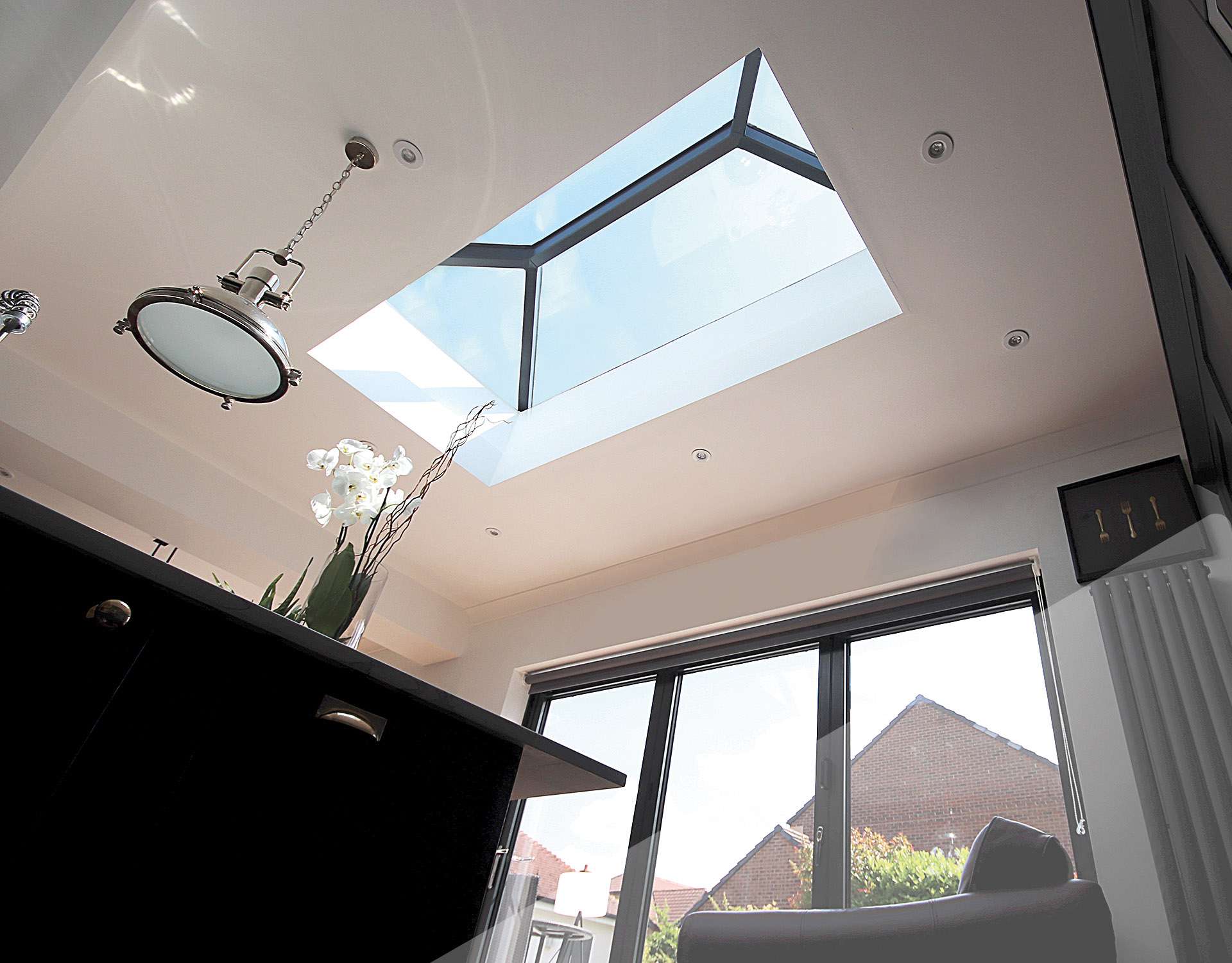 A worms eye view of a black Korniche roof lantern in a modern kitchen furnished with black counter tops a brown sofa chair and dark grey radiator. A rustic styled light hangs from the ceiling.