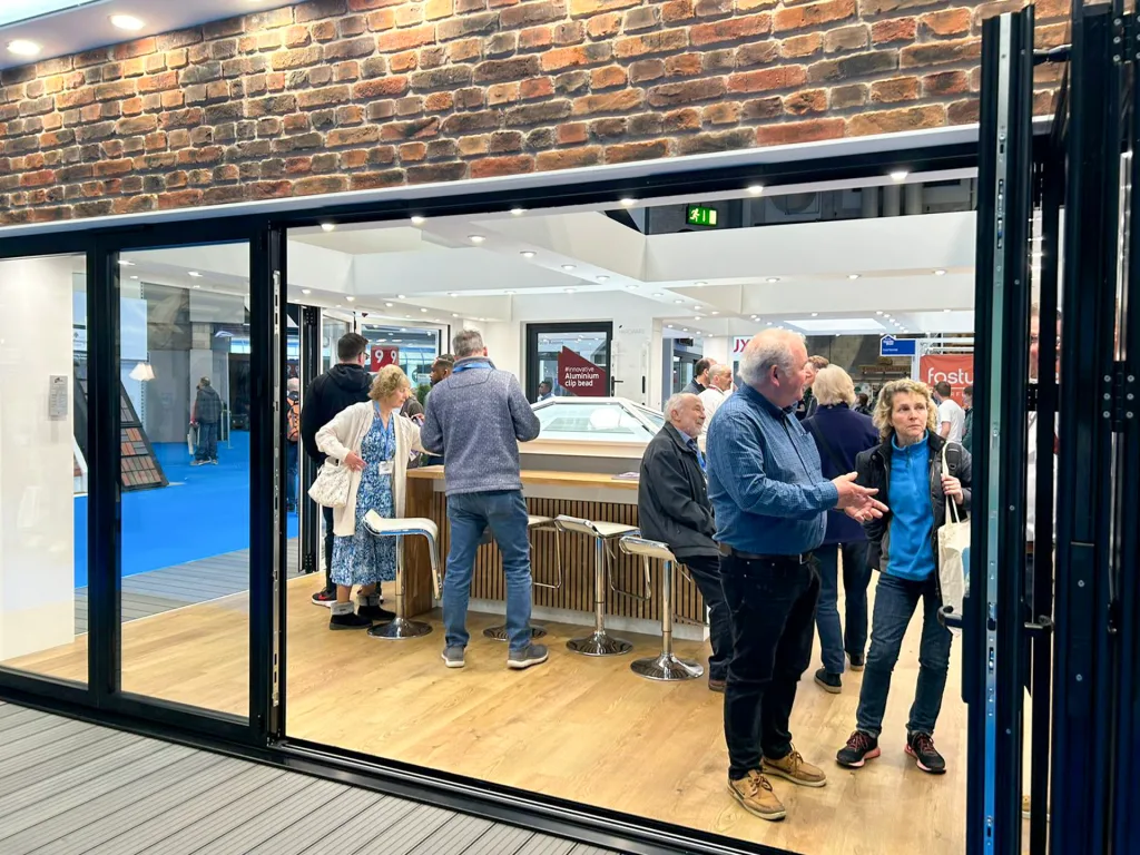 A view through a Korniche bi-folding door showcases a roof lantern in the center of the stand. Attendees gather around the lantern, examining it while conversing with a friendly Korniche team member.