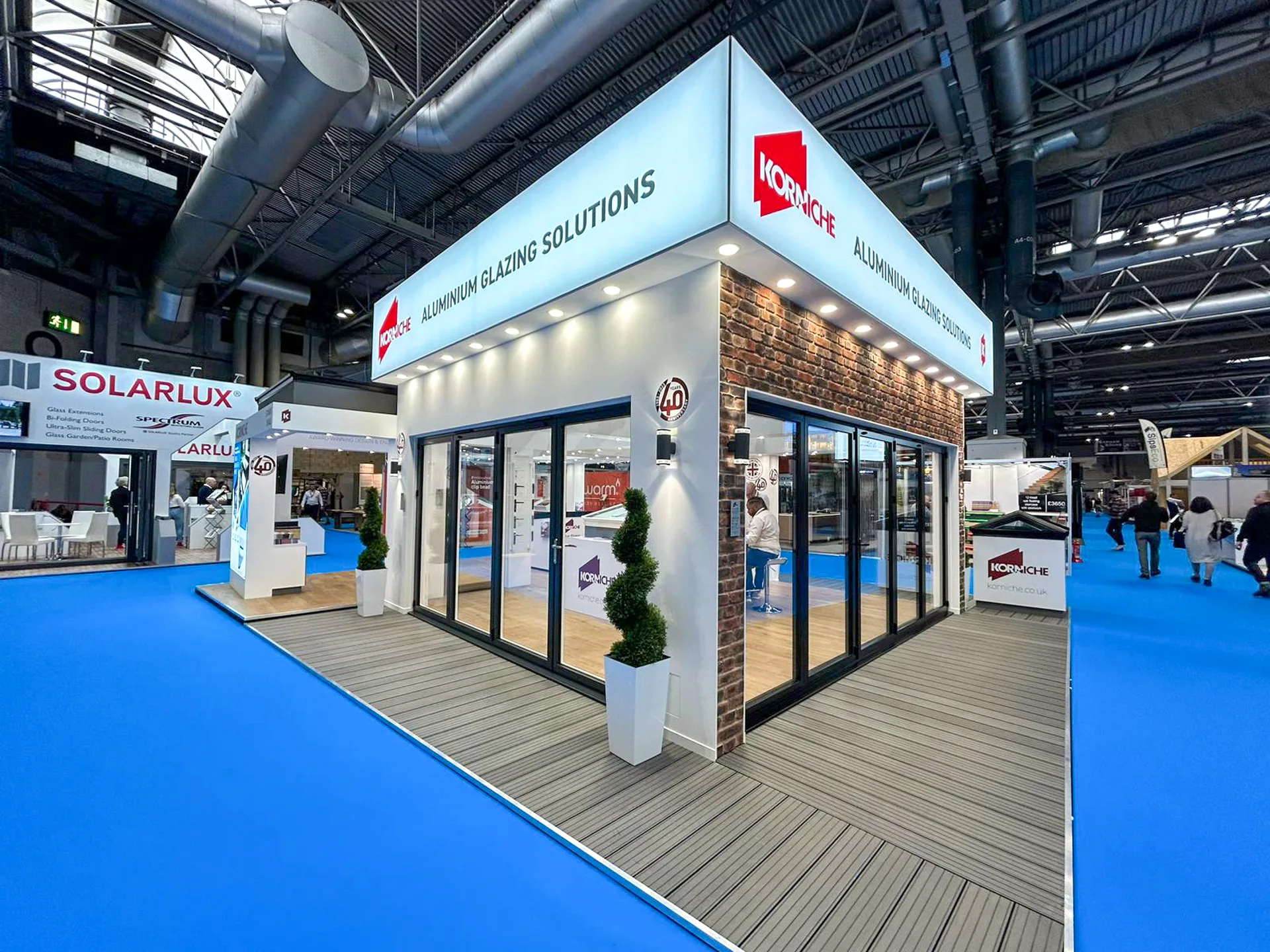 A wide shot of the National Homebuilding & Renovating Show floor before the show begins. The Korniche stand occupies a prominent location, showcasing bi-folding doors, roof lanterns, and a Flat Glass Rooflight System.