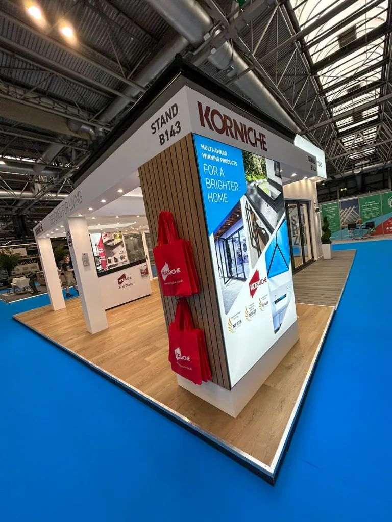 The corner of the Korniche stand at the National Homebuilding & Renovating Show. It features the Korniche logo, stand number B143, a display of Korniche tote bags, and a lit sign showcasing their award-winning products.